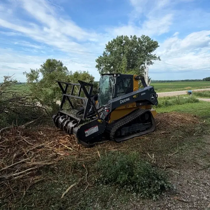Skid steer with a mulching head working beside a brush pile on a gravel road